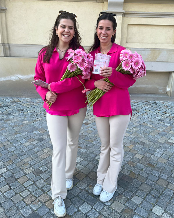 Zwei Frauen in pinken Pullovern halten Blumen in den Armen und haben Flyer in der Hand.